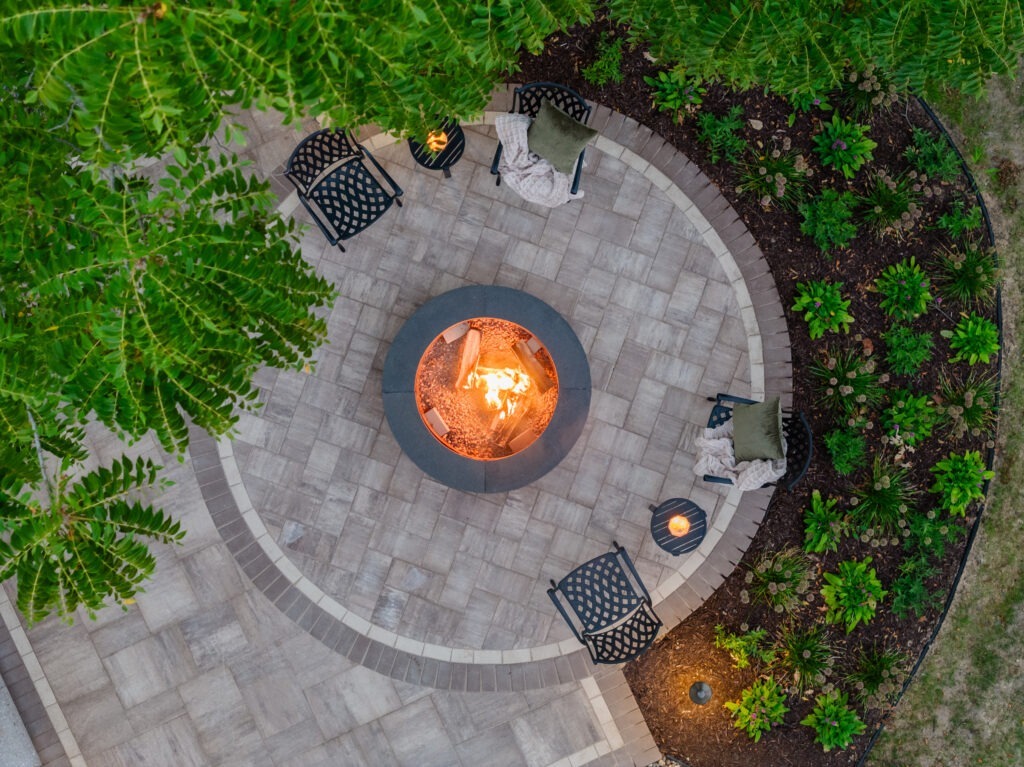 Overhead drone view of a custom paver patio with a glowing firepit in Hudson, WI, showcasing expert landscape design.