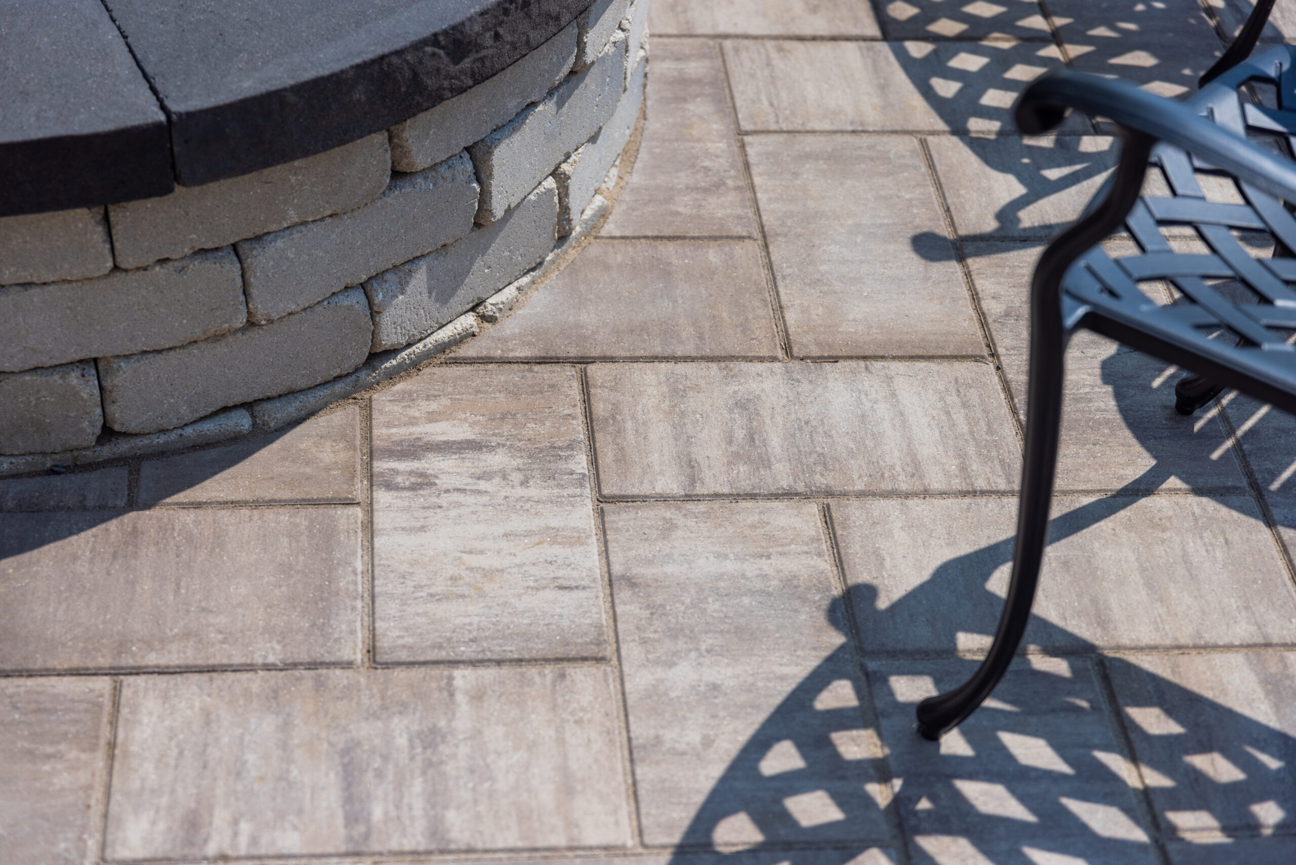Close-up of paver patio in Hudson, WI, showing the edge of a raised firepit with a chair casting a shadow.