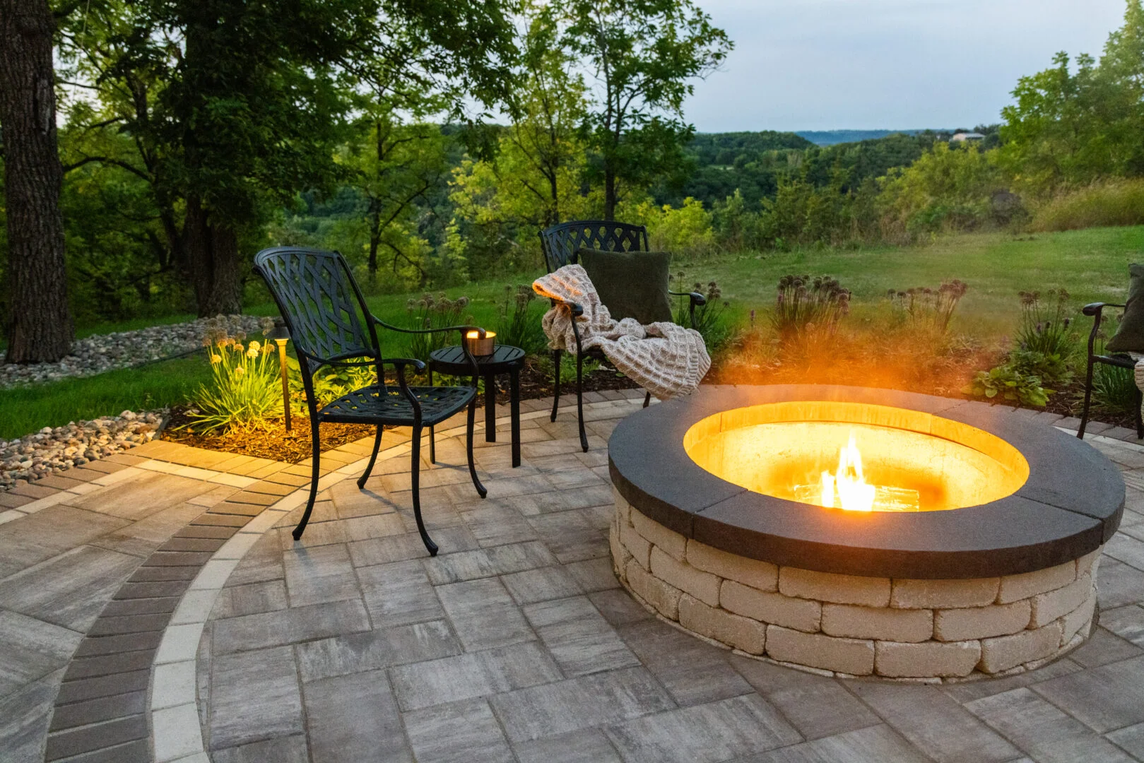 Raised wood-burning firepit on a paver patio in Hudson, WI, with cozy furniture, lush plants, and landscape lighting at dusk.